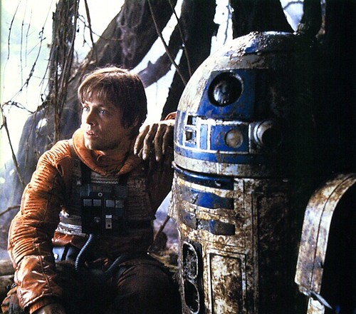Scene from Empire Strikes Back: Luke  Skywalker in orange flightsuit is sitting next to R2D2. Both are muddy in the aftermath of the droid's near-ingestion by swamp shark.