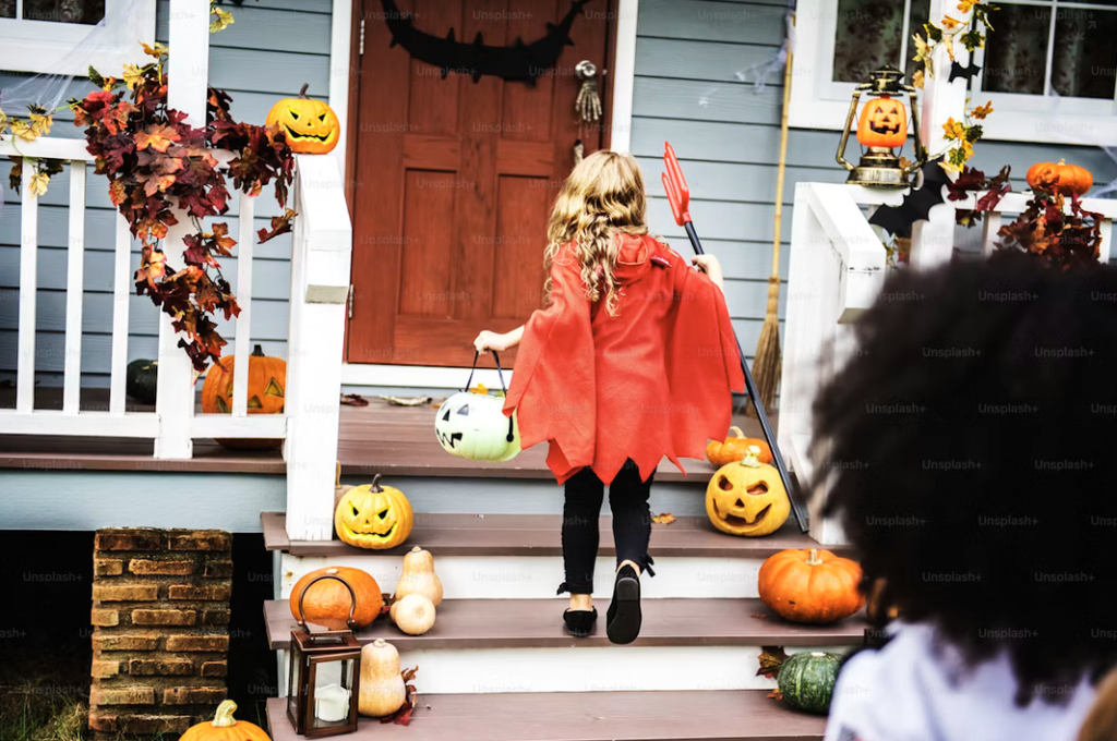 Photograph of a porch decorated for Halloween with jack-o-lanterns, leaf garlands, and bats. In the center of the image, a blond girl in a red and black devil costume carrying a pitchfork and a treat bucket ascends the stairs. The dark curly head of a second child is visible in the foreground right bottom corner. 